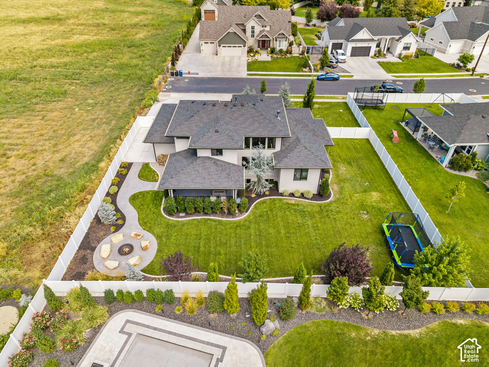 Expansive aerial view showcasing a modern two-story residence with a dark-pitched roof, situated on a professionally landscaped lot featuring manicured lawn, curved concrete driveway, mature plantings including ornamental trees and shrubs, circular hardsc
