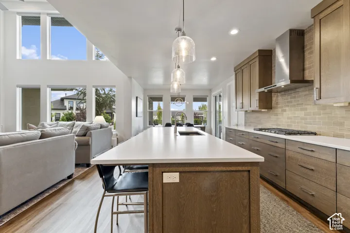 Open-concept kitchen featuring an expansive center island, integrated gas range with stainless steel hood, wood-finish cabinetry, and a tile backsplash
