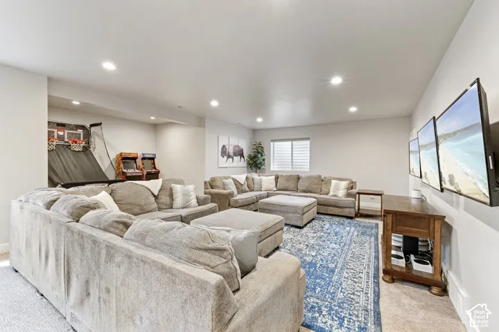 Expansive recreational room featuring a sectional layout with abundant seating, recessed ceiling lighting, wood-finish flooring, a blue area rug, wall-mounted flat-screen television, and large windows with plantation shutters flooding the space with natur