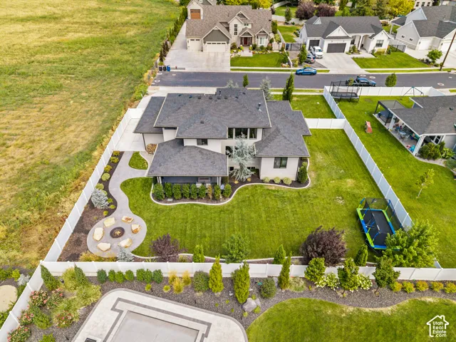 Expansive aerial view showcasing a modern two-story residence with a dark-pitched roof, situated on a professionally landscaped lot featuring manicured lawn, curved concrete driveway, mature plantings including ornamental trees and shrubs, circular hardsc