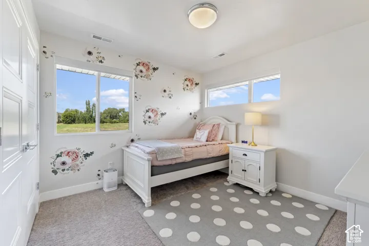 Bright bedroom featuring dual large windows with natural light, neutral wall finishes, hardwood-finish flooring, a patterned area rug, and a bedside nightstand with integrated lighting