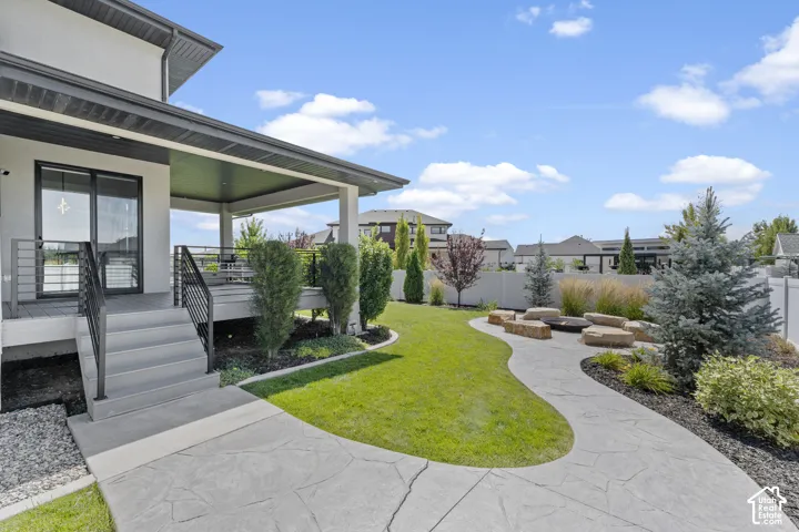Expansive backyard oasis featuring a manicured lawn, curved concrete patio with integrated walkways, mature evergreen landscaping, and a modern covered patio structure with glass sliding doors extending from the residence