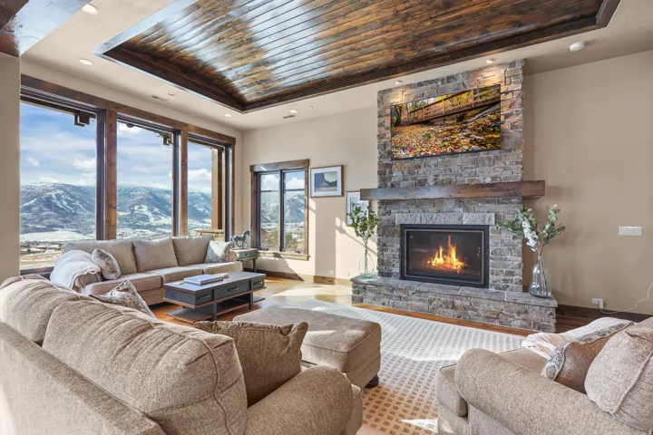 Recessed wood-panel ceiling, stone-clad fireplace with wood mantel, large picture windows, and wood-finish flooring