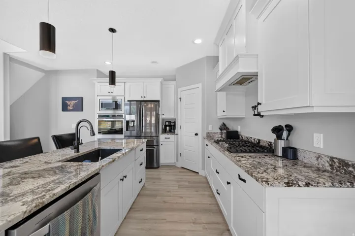 White shaker cabinetry, stainless steel appliances, wood-finish flooring, island with integrated sink, and stone countertops