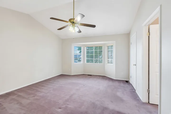 Bright corner bedroom showcasing vaulted ceilings, ceiling fan with integrated lighting, and triple-window configuration allowing abundant natural light