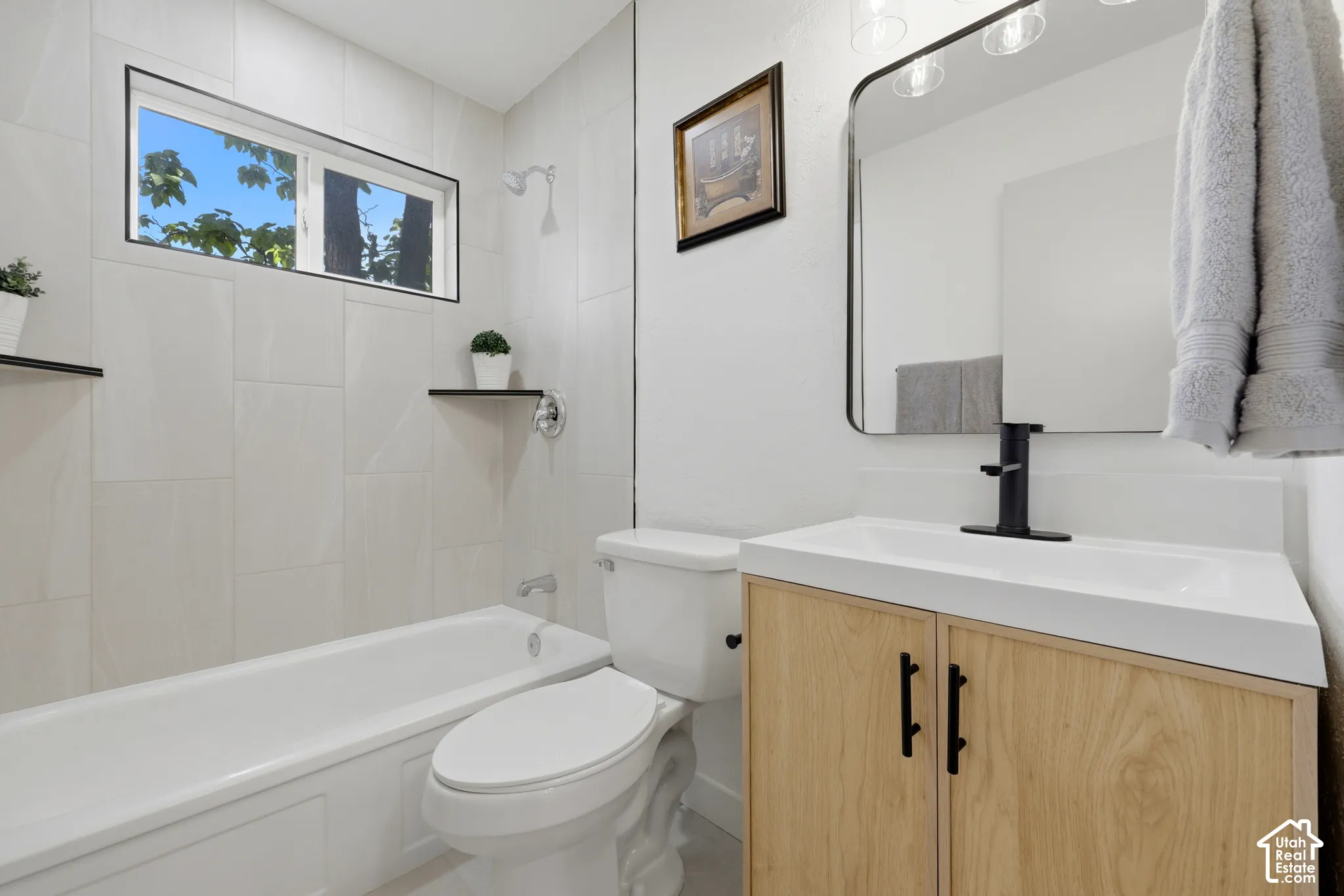 White tile shower surround with integrated tub, natural wood-finish vanity with white countertop and integrated sink, black-framed mirror, and black wall-mounted faucet