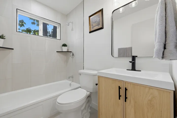 White tile shower surround with integrated tub, natural wood-finish vanity with white countertop and integrated sink, black-framed mirror, and black wall-mounted faucet