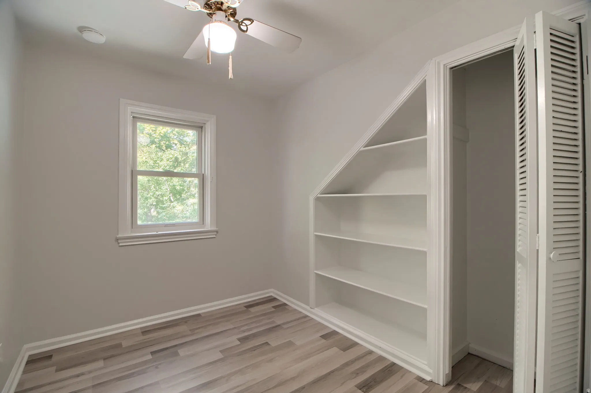 Recessed built-in shelving, wood-finish flooring, single window, louvered bi-fold closet doors, and a ceiling fan with integrated lighting