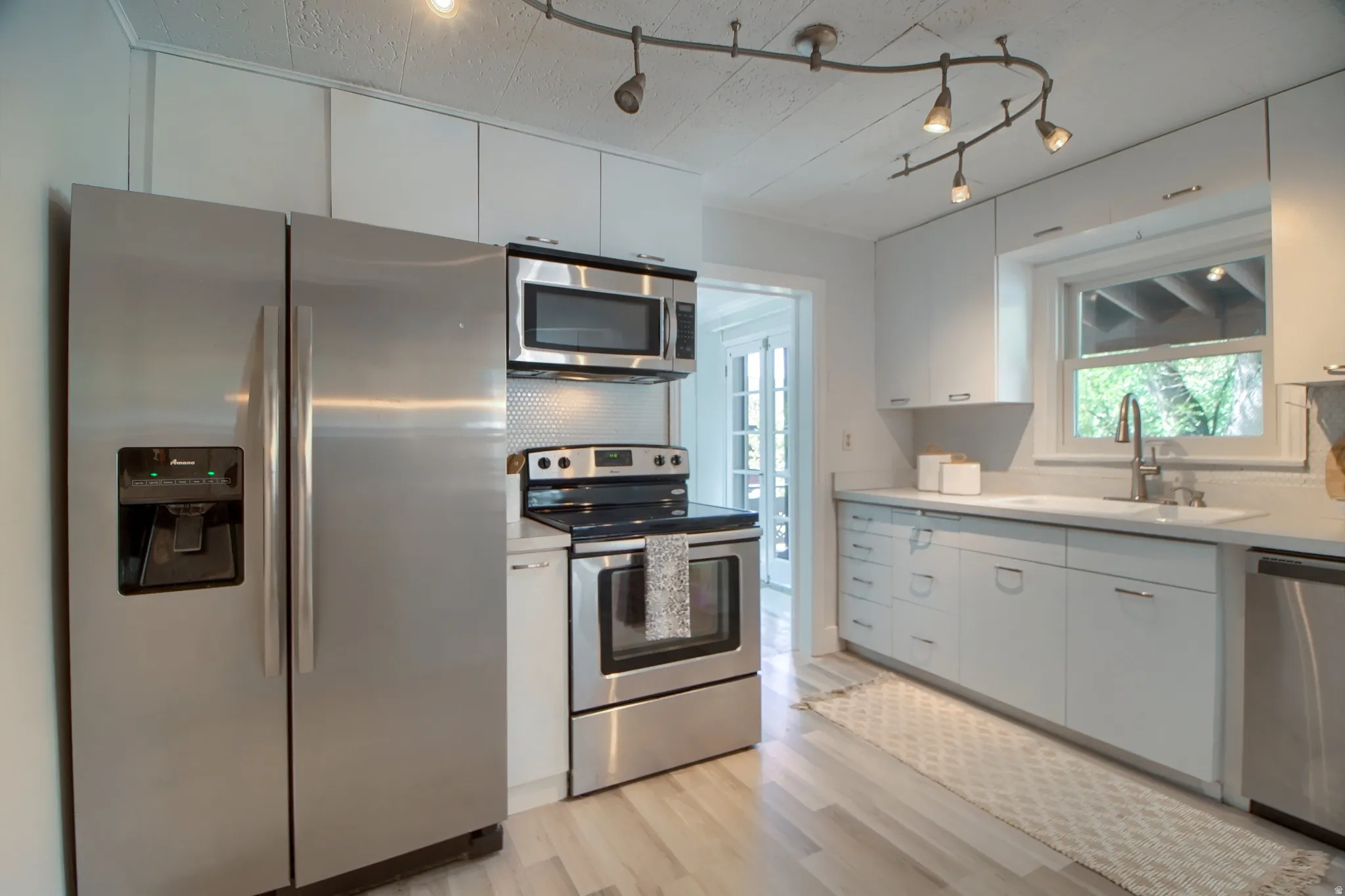 Stainless steel appliances, white flat-panel cabinetry, light wood-finish flooring, white countertop, and a track lighting system