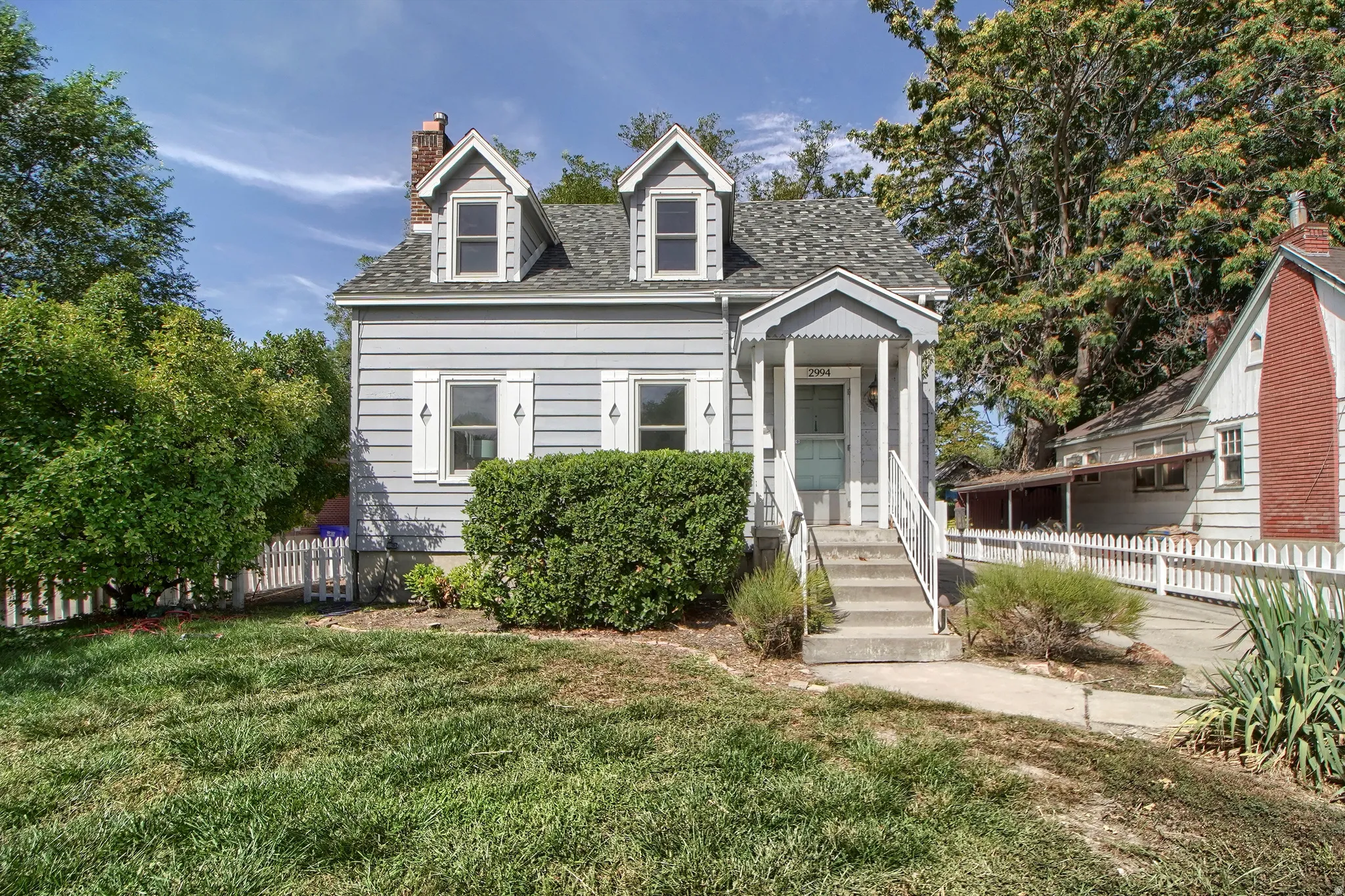 Light blue siding exterior with white trim, a covered porch entrance, multiple dormer windows, and a picket fence