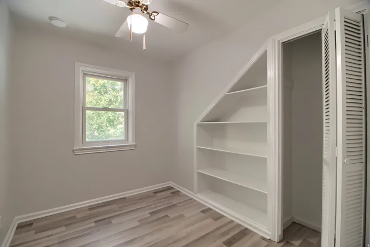 Recessed built-in shelving, wood-finish flooring, single window, louvered bi-fold closet doors, and a ceiling fan with integrated lighting