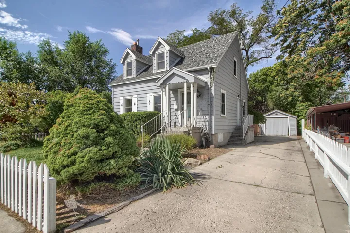 Gabled roofline with dormer windows, covered front porch, picket fence, detached garage, and mature landscaping