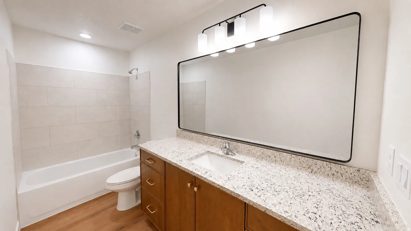 Wood-finish flooring, white bathtub with a tiled surround, wood vanity with granite countertop, integrated sink, and large wall-mounted mirror