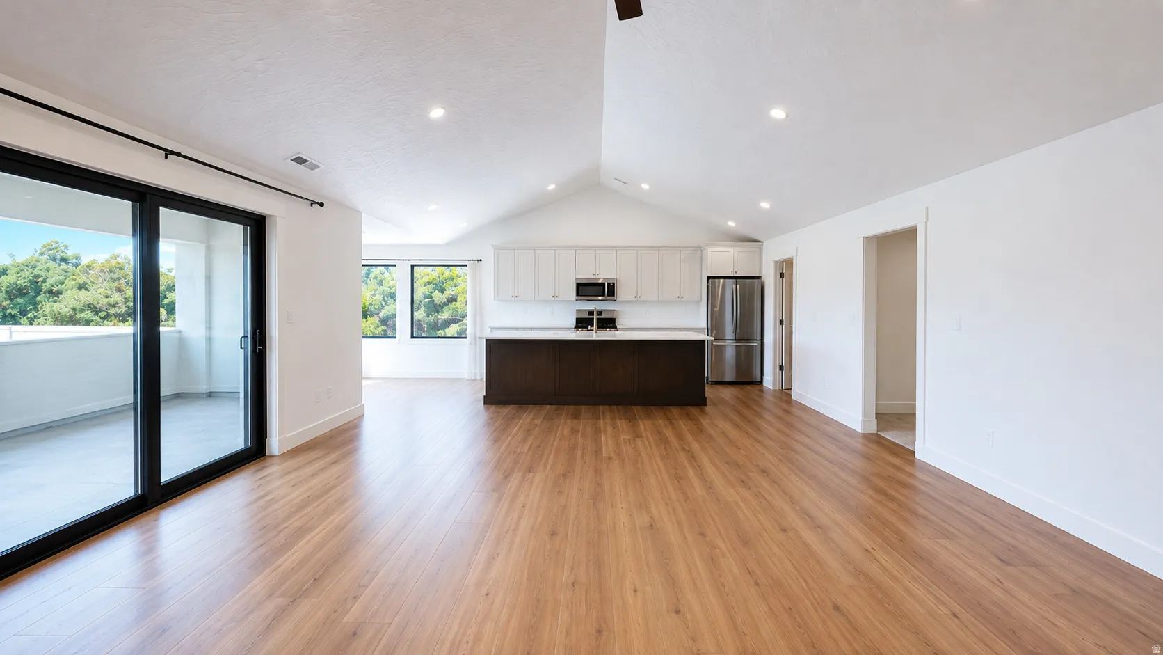 Spacious living area featuring wood-finish flooring, vaulted ceilings, recessed lighting, and a kitchen with white upper cabinetry, dark lower cabinetry, and stainless steel appliances