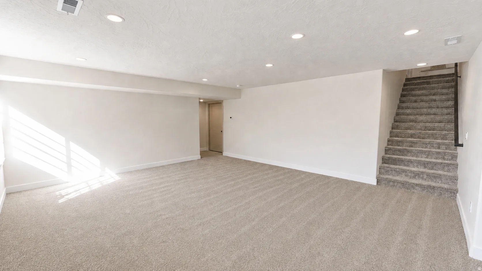 Recessed ceiling lighting, neutral carpet flooring, light-toned wall finishes, and a carpeted staircase with a dark handrail