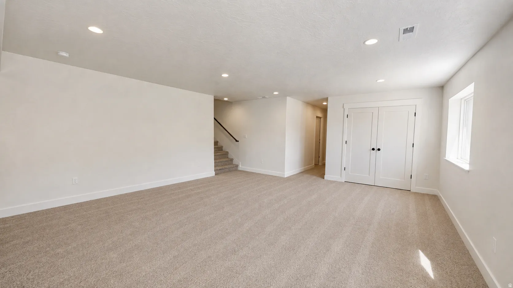 Recessed ceiling lighting, light-colored carpeting, white double doors, white window trim, and a visible staircase