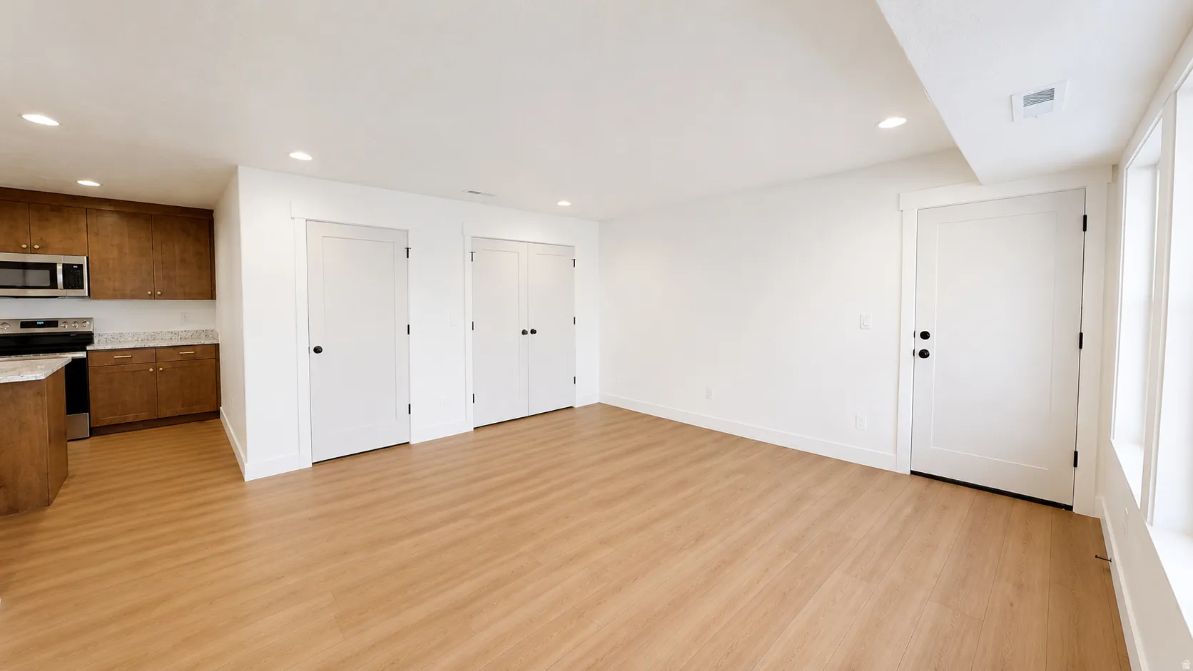 Wood-finish flooring throughout, recessed lighting, white walls, white trim, and a kitchen area featuring wood cabinetry, stainless steel appliances, and a light-colored countertop