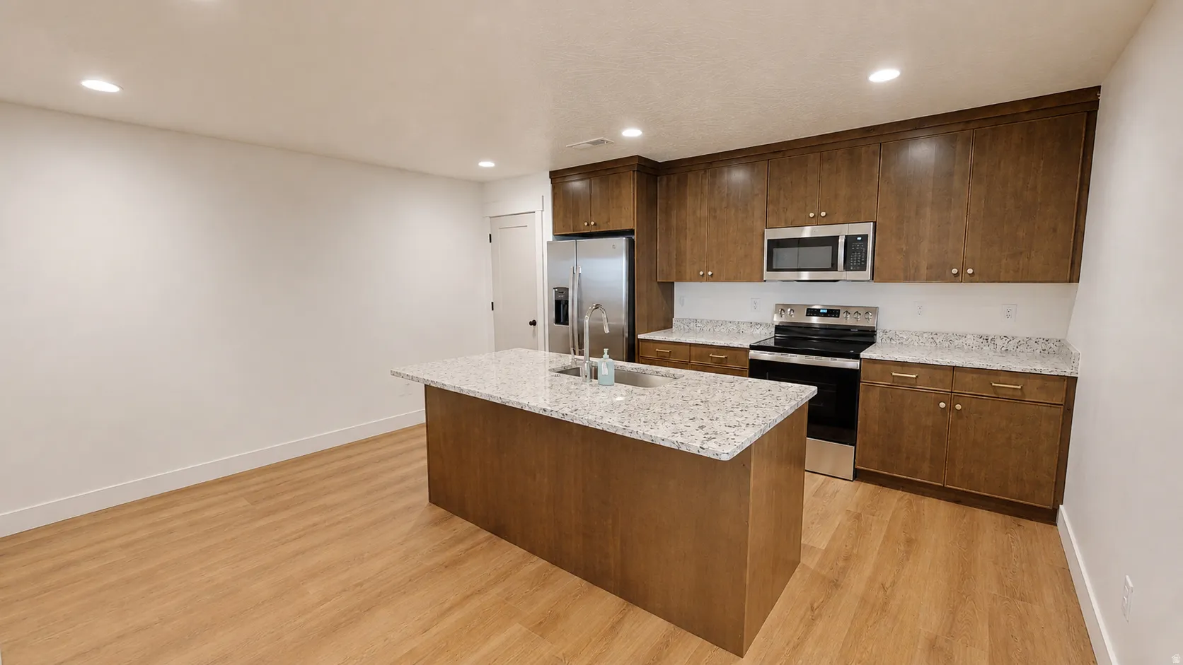 Kitchen featuring wood-finish flooring, wood cabinetry with gold-tone hardware, a center island with a sink, and stainless steel appliances