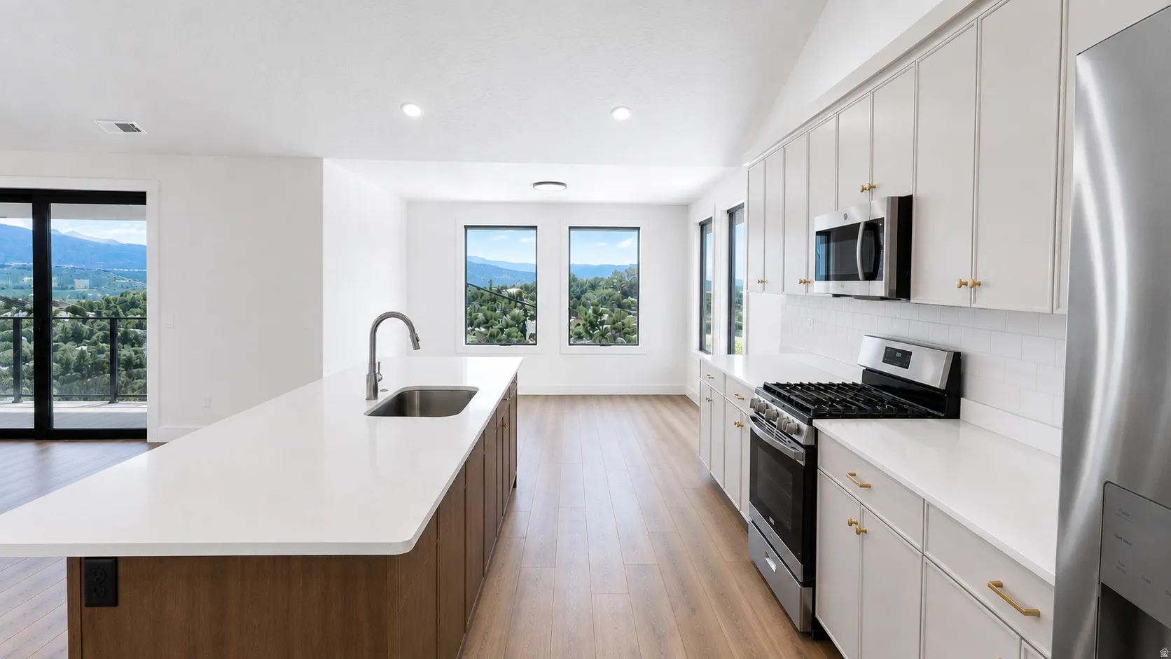 Kitchen featuring wood-finish flooring, an expansive island with an undermount sink, white cabinetry with gold-toned hardware, and stainless steel appliances