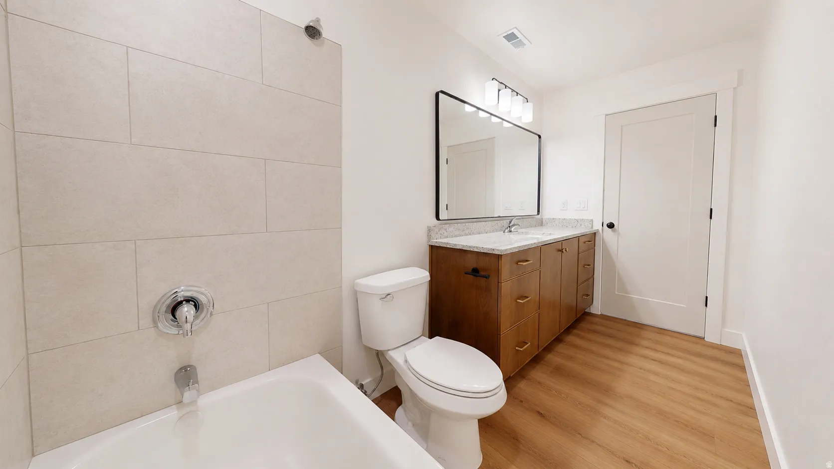 Wood-finish flooring, single vanity with light-colored countertop, framed mirror, and a full bathtub with light-toned tiled walls