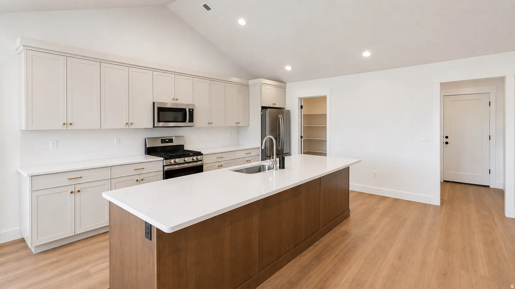 Vaulted ceiling with recessed lighting, light-toned cabinetry, stainless steel appliances, central island with sink, and wood-finish flooring