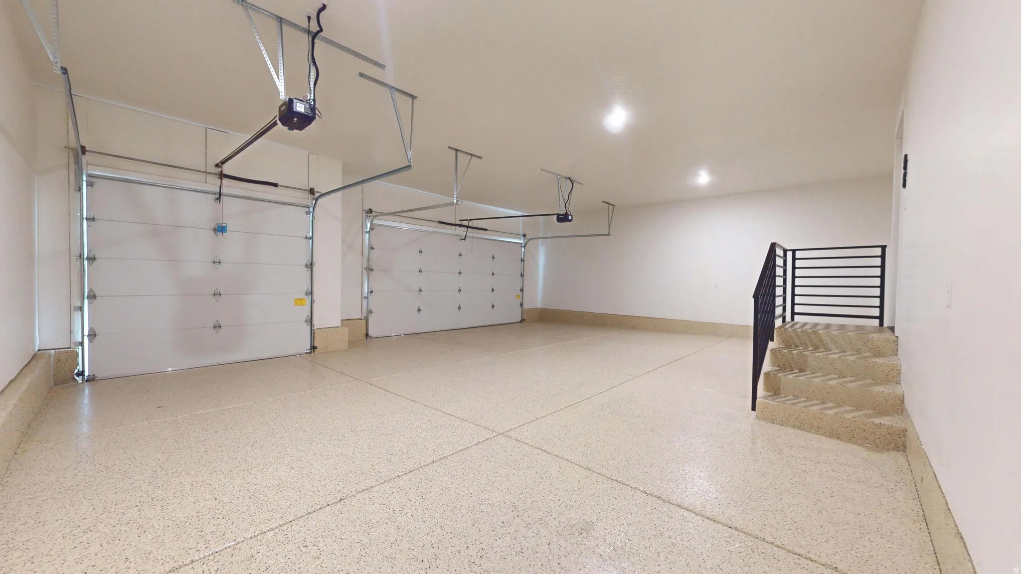 Two automatic garage doors, speckled epoxy flooring, recessed lighting, and a staircase with a black metal railing