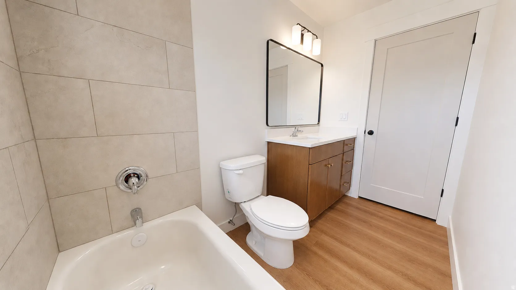 White bathtub with chrome fixtures, tiled shower surround, wood-finish flooring, wood vanity with a white countertop, and a rectangular mirror with a three-light fixture
