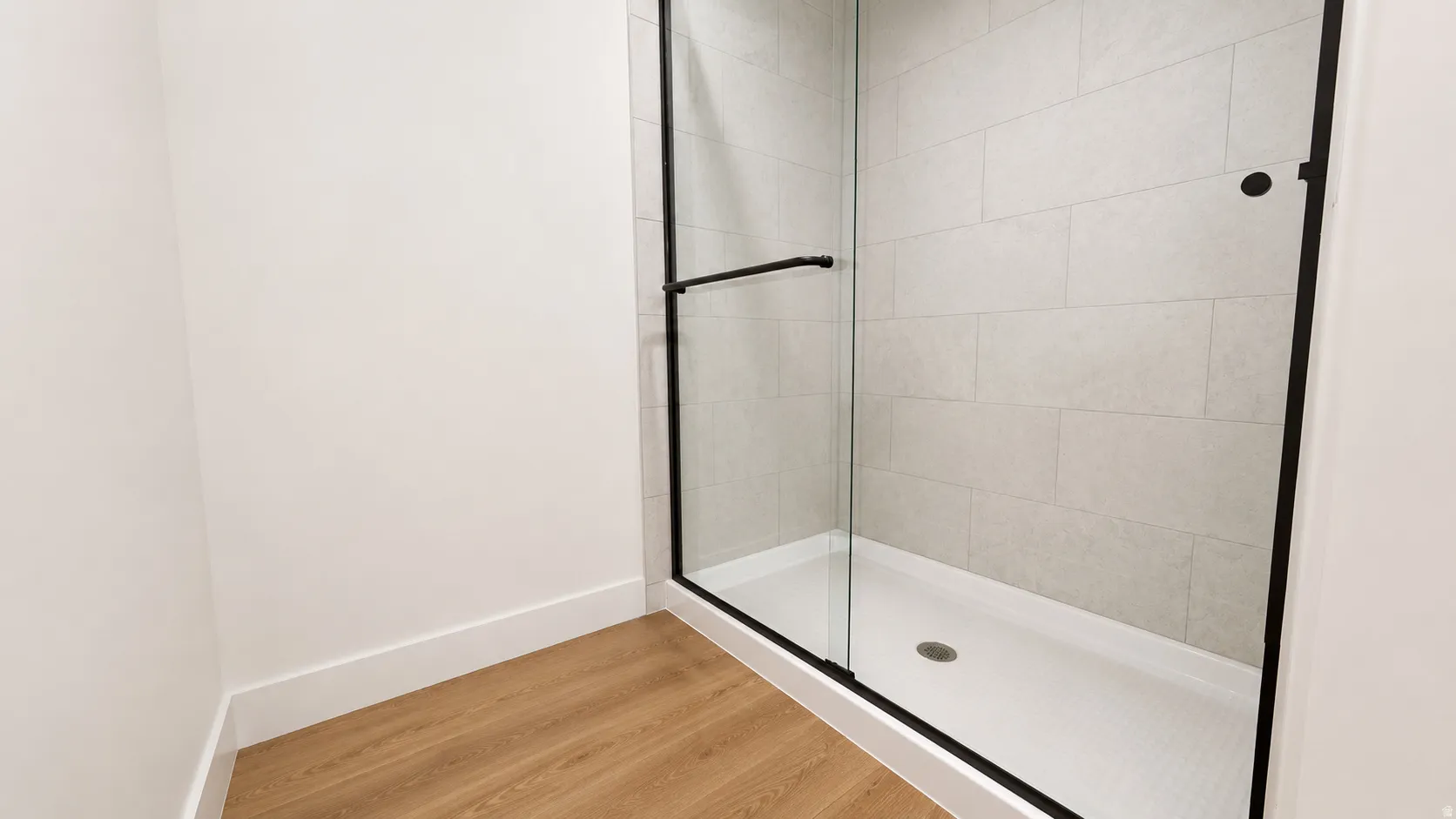 Wood-finish flooring, white wall paint, and a shower enclosure featuring a clear glass door, matte black hardware, and light-toned tile surround