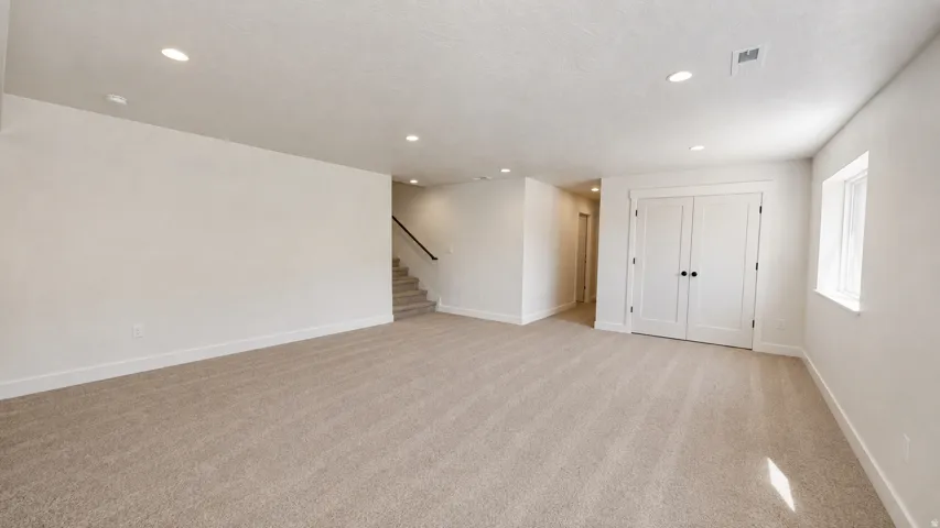 Recessed ceiling lighting, light-colored carpeting, white double doors, white window trim, and a visible staircase