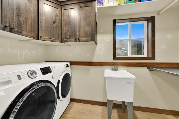 Wood-finish cabinetry, a utility sink with dual-handle faucet, a window with wood trim, wood-finish flooring, and a built-in shelf