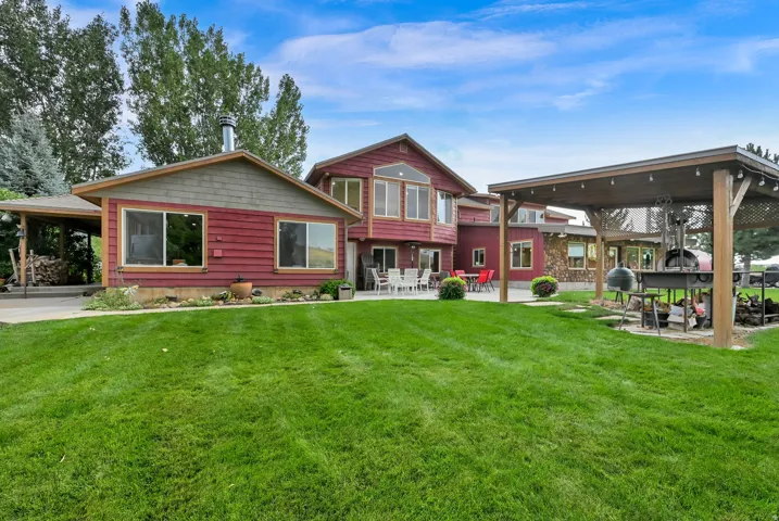 Expansive green lawn, multi-level red siding exterior, covered patio, outdoor cooking area, and mature trees