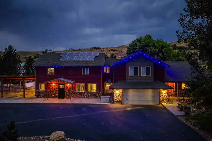 Modern residence showcasing a striking red exterior with professional landscape lighting, solar panel array on the roofline, attached two-car garage with illuminated entry, and mature landscaping with established trees framing the property