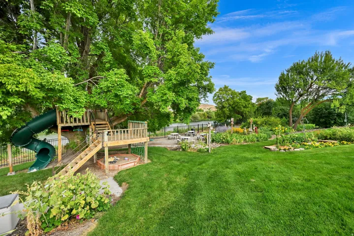 Expansive backyard with a manicured lawn, established garden beds, mature trees, and a built-in wooden play structure with a slide and sandpit