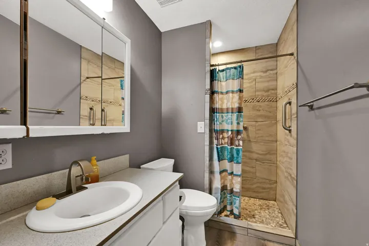 Vanity with an integrated sink and laminate countertop, framed medicine cabinet, toilet, and a tiled shower with a pebble floor