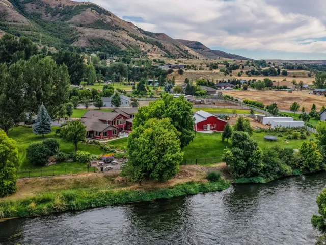 Riverfront property featuring a red-sided residence with green landscaping, mature trees, and mountainous terrain in the background