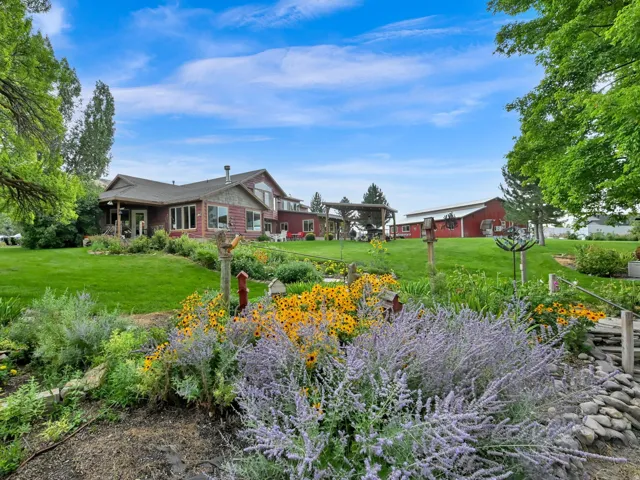 Expansive lawn with mature trees, colorful garden beds, and a red-sided residence featuring multiple windows and a covered patio