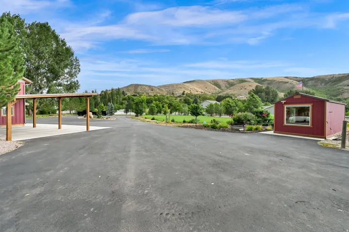 Extensive paved driveway, covered carport, detached red outbuilding with a window, landscaped area, and distant hills