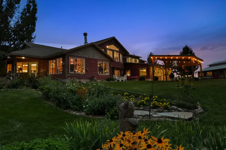 Expansive lawn area with a multi-level wood-sided exterior, numerous windows, covered patio with string lighting, and established garden beds