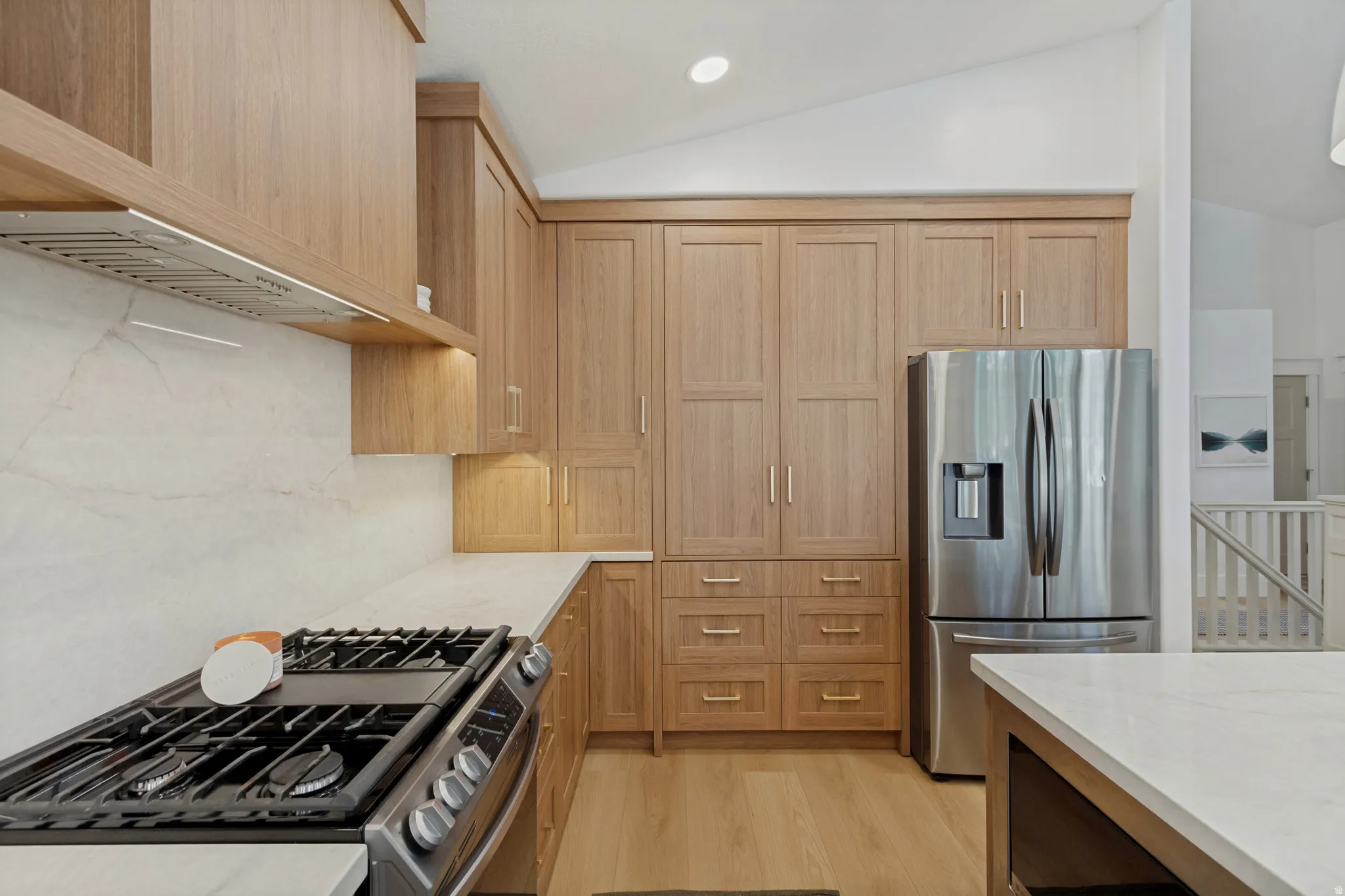 Integrated stainless steel refrigerator, gas range with coordinating ventilation hood, extensive wood-finish cabinetry, light countertops, and wood-finish flooring