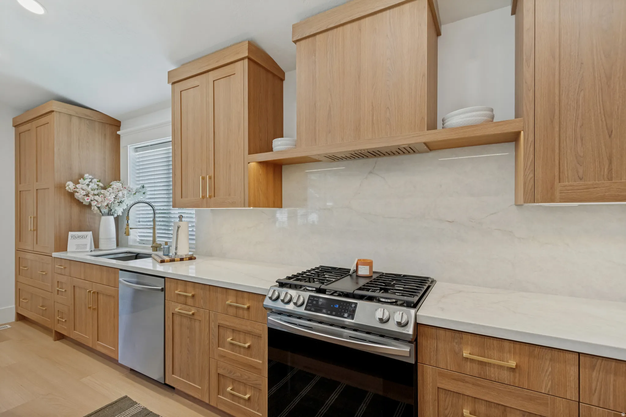 Wood-finish cabinetry with brass hardware, light-toned countertop, integrated stainless steel dishwasher, stainless steel range, and light-toned backsplash
