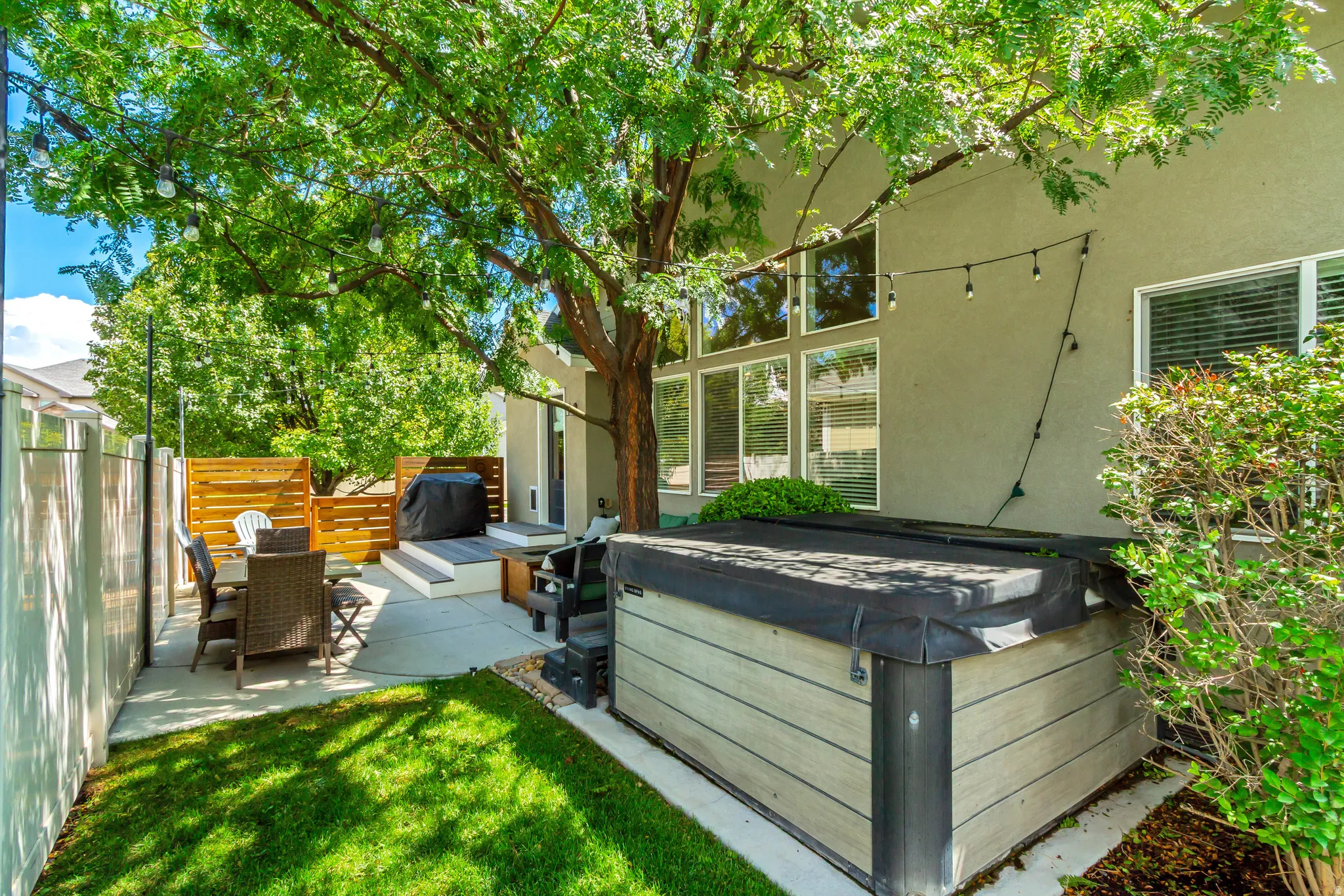 Mature shade trees create a serene backyard oasis featuring a manicured lawn, hot tub with integrated seating, wooden privacy fencing, and direct access to the home's sliding glass doors