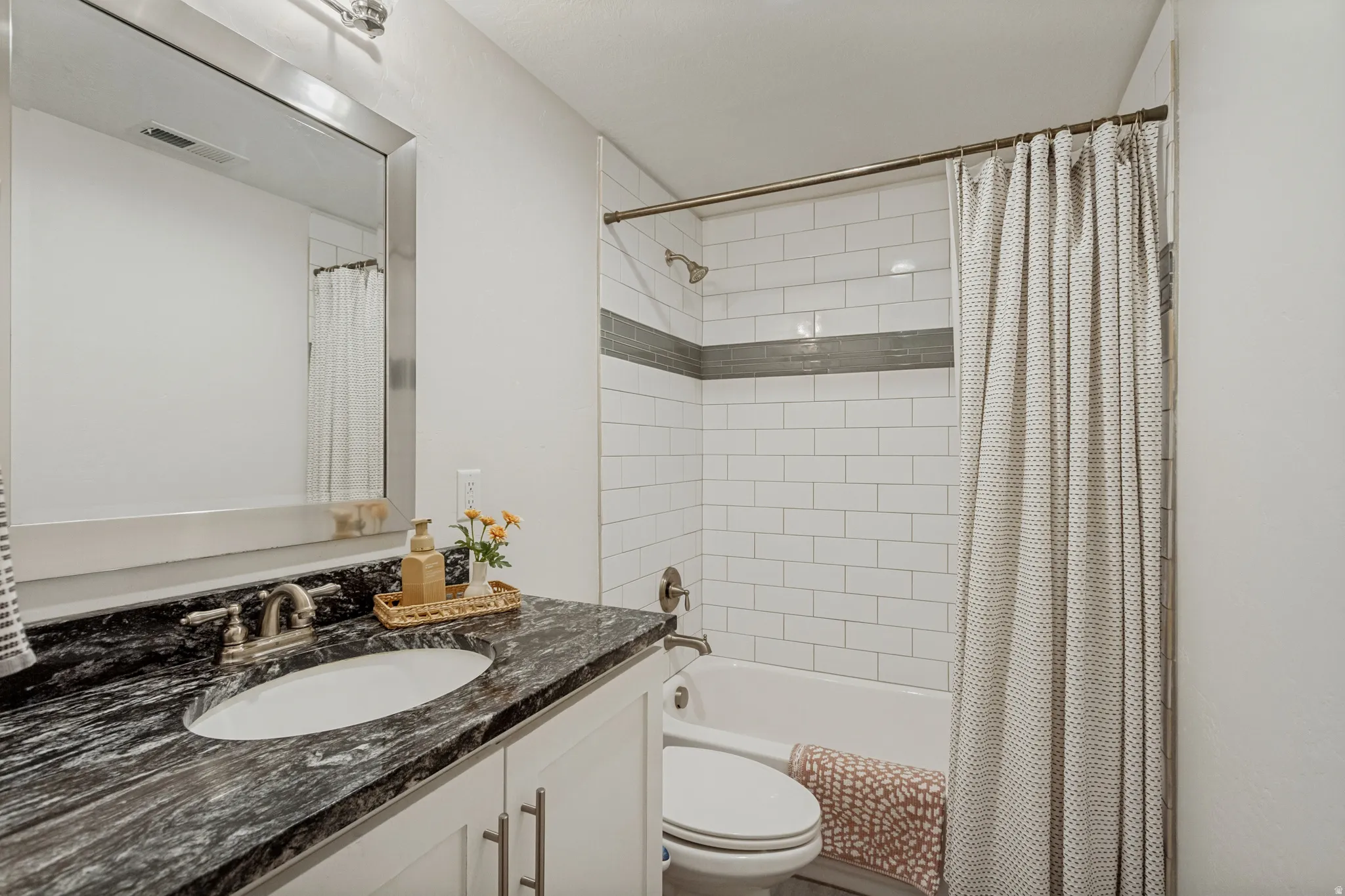 White subway tile shower surround with a gray accent band, and a vanity featuring a dark patterned countertop with an oval undermount sink and brushed nickel faucet