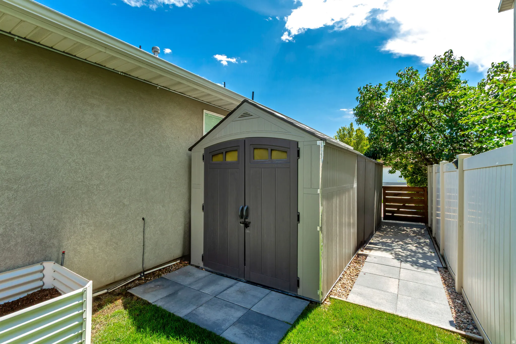 Detached outdoor storage shed with dual-paneled doors, a paver stone landing, a gated side path, and a privacy fence