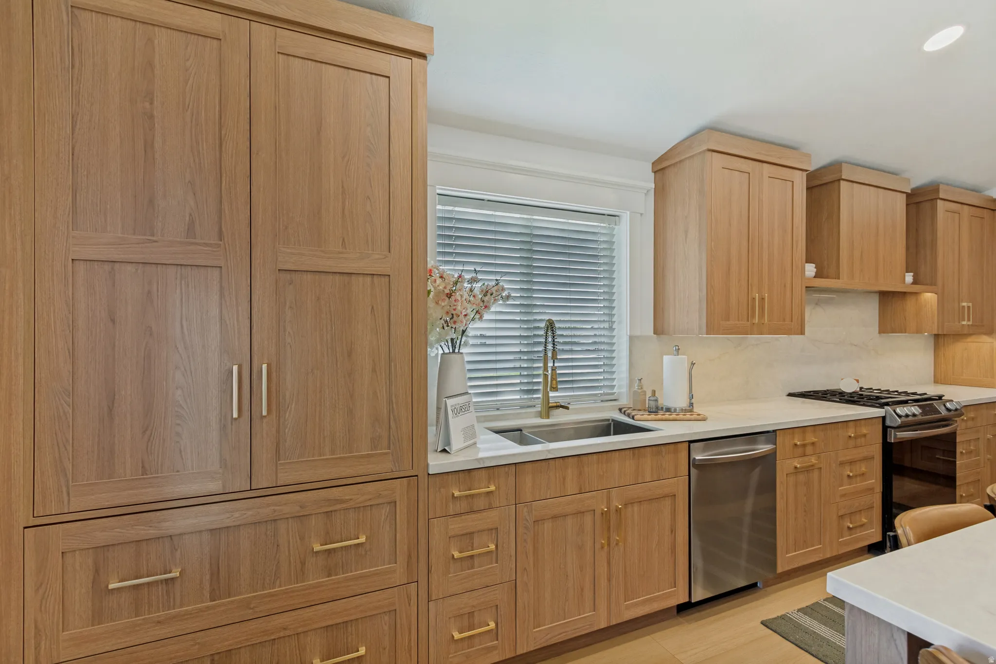Wood-finish cabinetry with brass hardware, dual basin stainless steel sink, stainless steel dishwasher, white countertops, and white tile backsplash