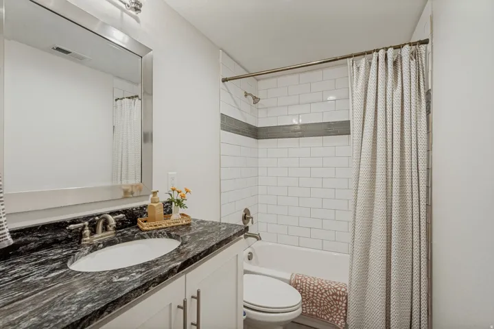 White subway tile shower surround with a gray accent band, and a vanity featuring a dark patterned countertop with an oval undermount sink and brushed nickel faucet