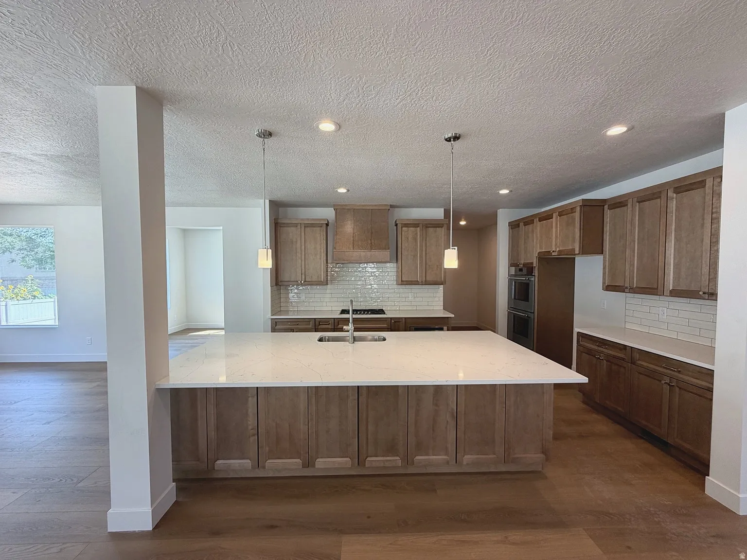 Spacious kitchen featuring a large island with sink, wood-finish cabinetry, subway tile backsplash, and stainless steel double ovens