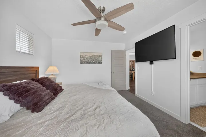 Wood-finish headboard, window with horizontal blinds, ceiling fan with light fixture, wall-mounted television, and carpet flooring