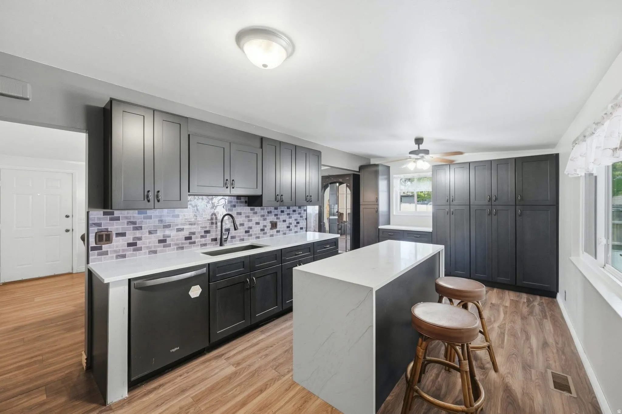 Kitchen featuring extensive dark cabinetry, light-toned countertops, a central island with waterfall edge, wood-finish flooring, and a tiled backsplash