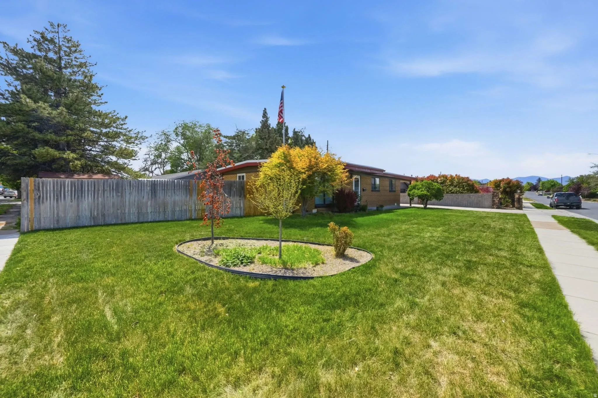 Expansive front lawn with a circular planting bed, mature trees, a wooden privacy fence, and a concrete sidewalk