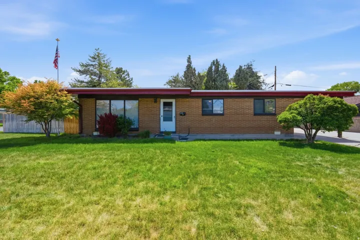 Brick ranch home featuring a sprawling manicured lawn, mature landscaping with established trees, multiple windows providing natural light, and a central entry with covered approach
